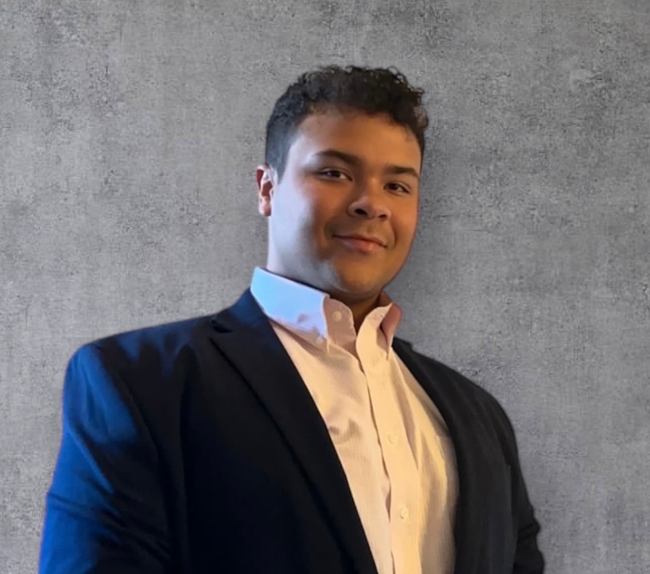 Young man with curly hair wearing a navy blazer and white shirt against grey wall.